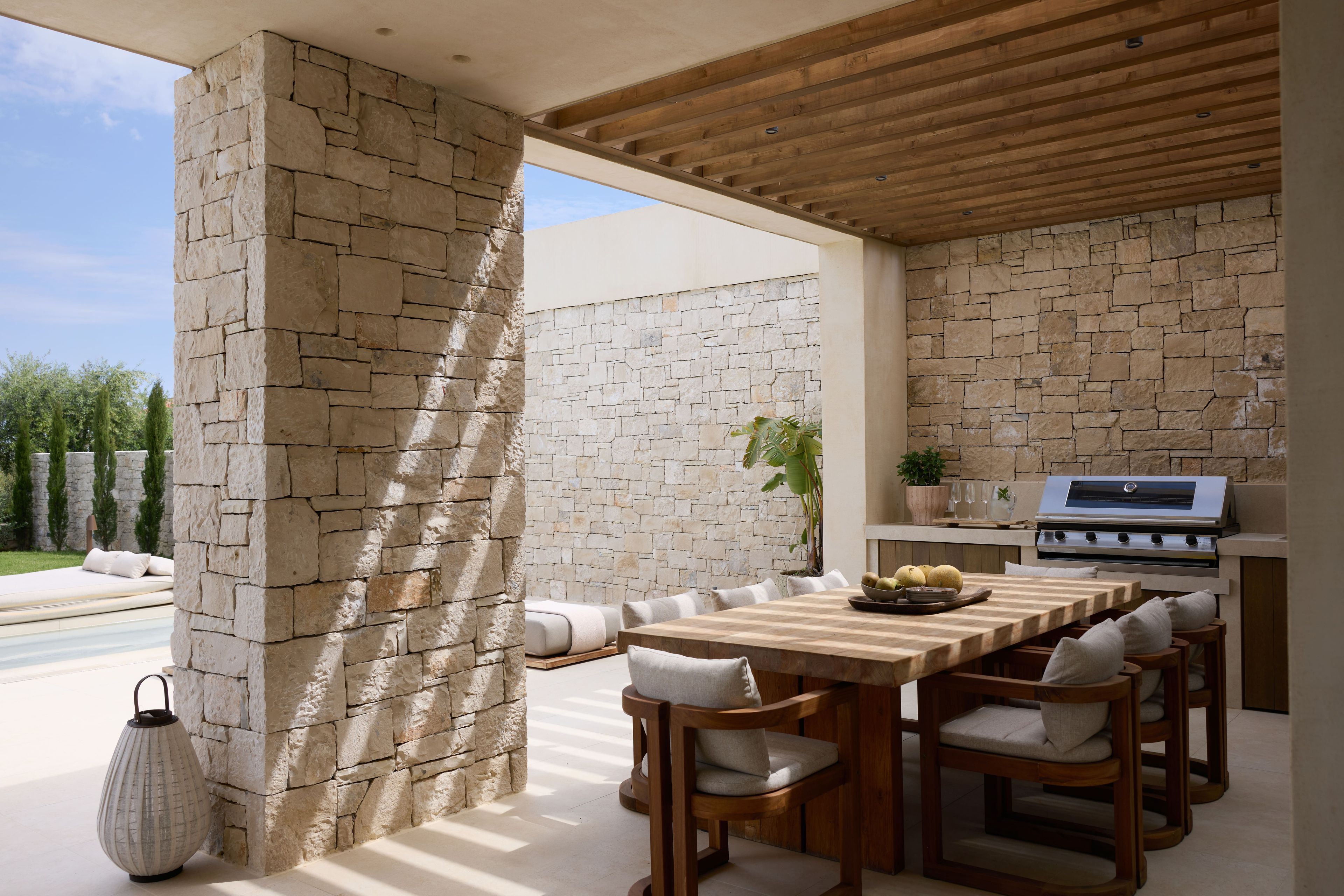 An outdoor patio with stone walls, a wooden ceiling, a dining table with chairs, and a grill.