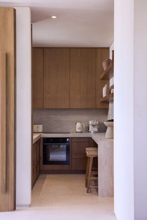 A compact kitchen with wooden cabinets, a built-in oven, a coffee machine, and a small seating area with stools.