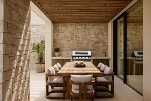 A patio with a stone wall, a wooden ceiling, a table with chairs, and a grill.