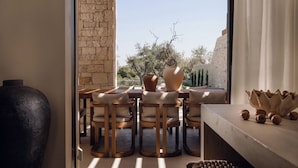 Un comedor con pared de piedra, sillas de madera, un jarrón grande y vistas a árboles y cielo.