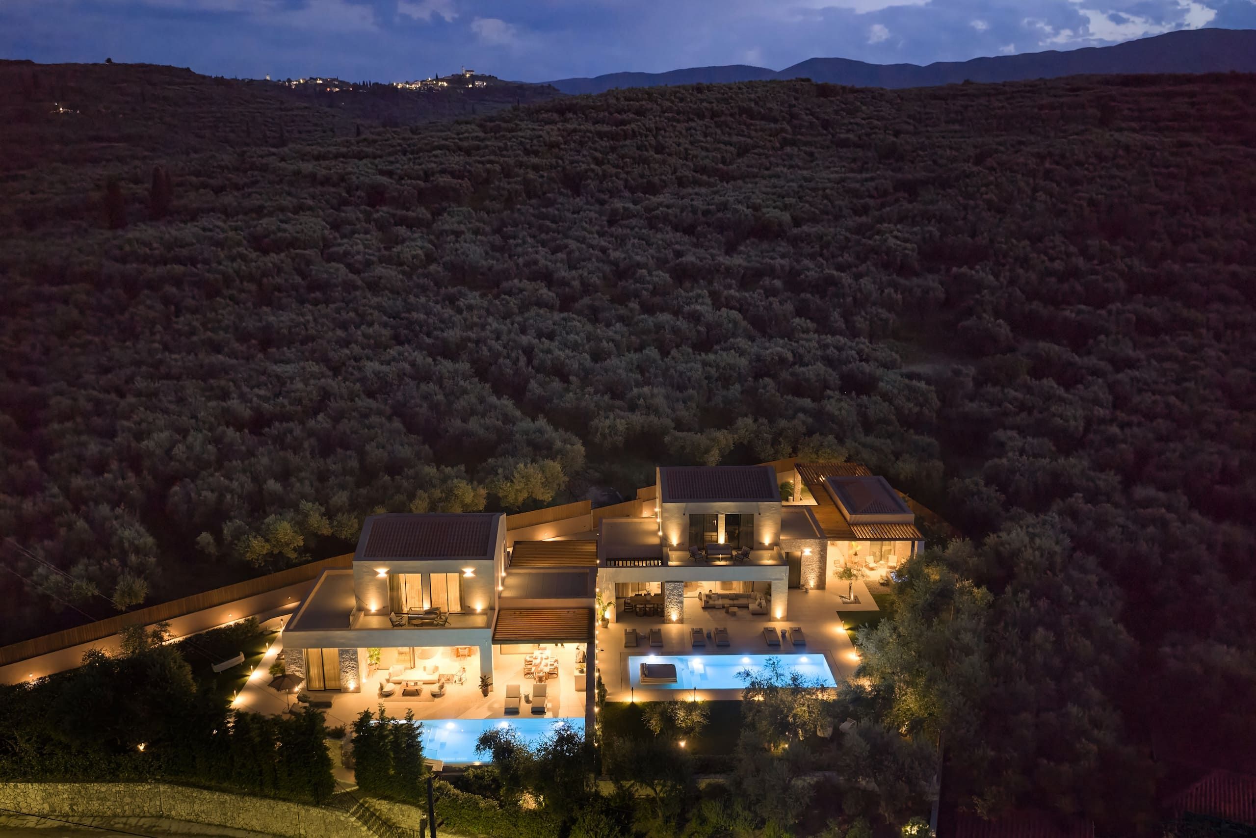 A modern two-story house with a swimming pool, surrounded by dense vegetation and hills.