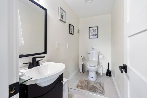 Une salle de bain moderne avec un lavabo blanc, un robinet noir et des toilettes. Des tableaux encadrés ornent le mur, et une pancarte indique : « Merci de ne pas vous asseoir pendant toute la durée du spectacle. »