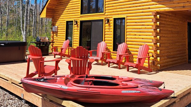 Una cabaña de troncos con un porche, sillas Adirondack rojas y un kayak rojo.