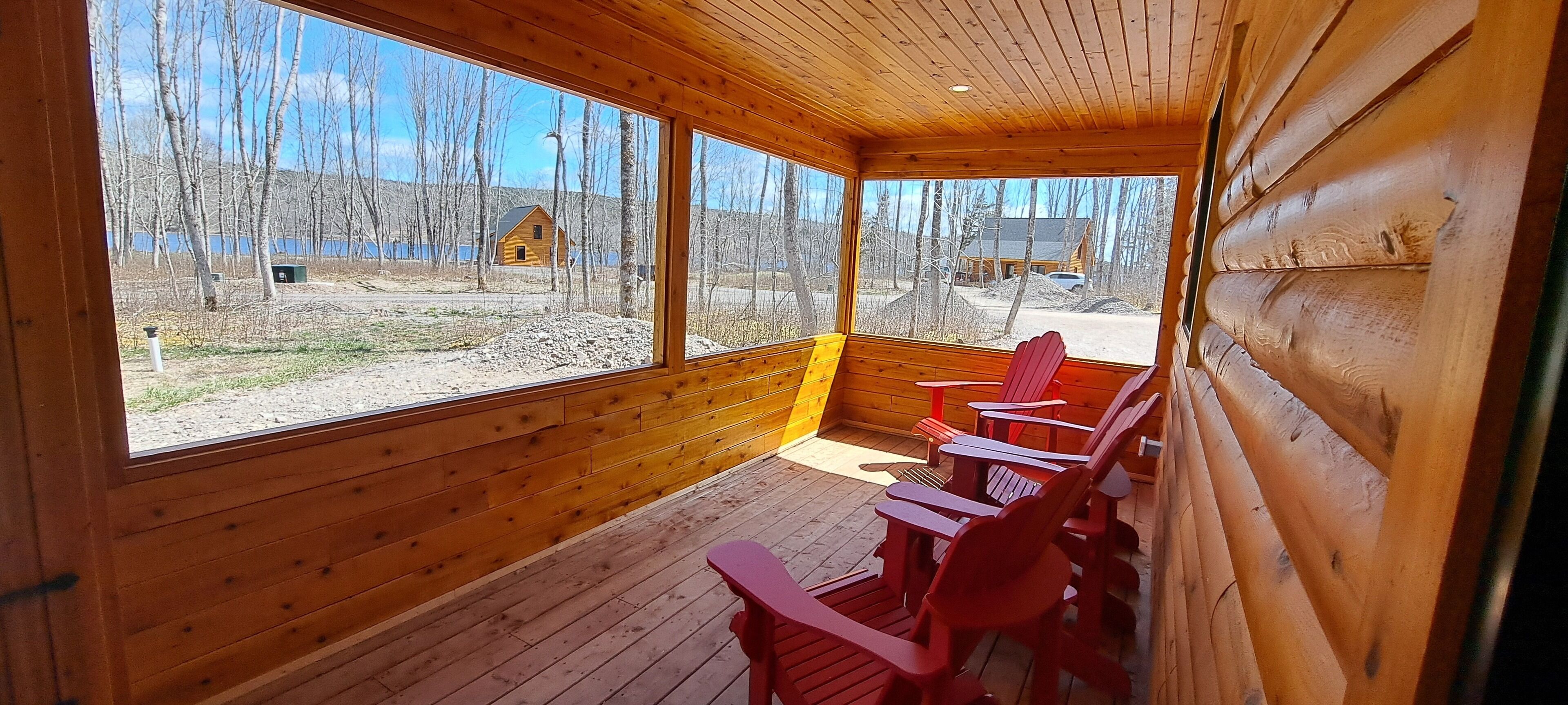 Eine Holzhütte mit großen Fenstern, roten Adirondack-Stühlen und Blick auf See und Wald.