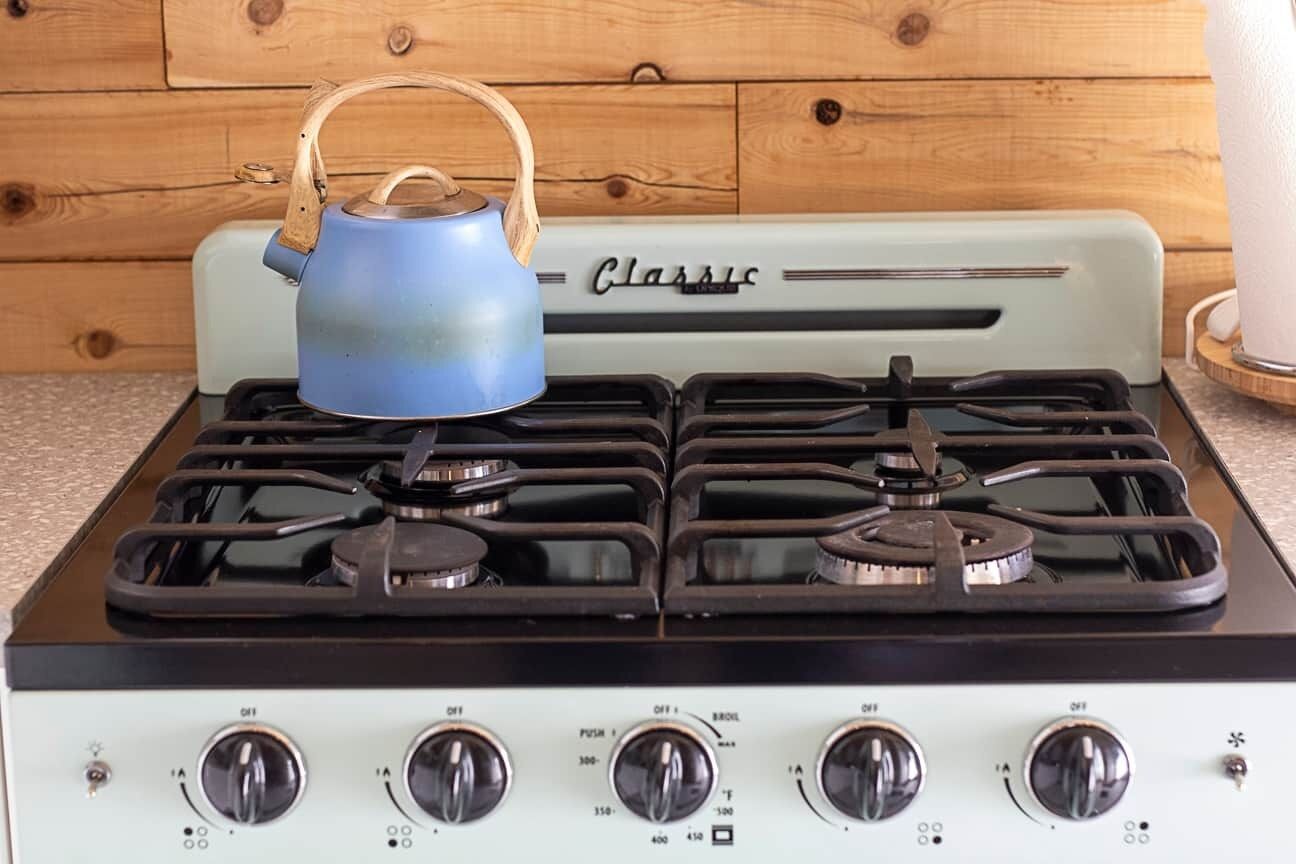 Une cuisinière ancienne, dotée de quatre brûleurs et surmontée d’une bouilloire bleue, le tout placé devant un meuble en bois.