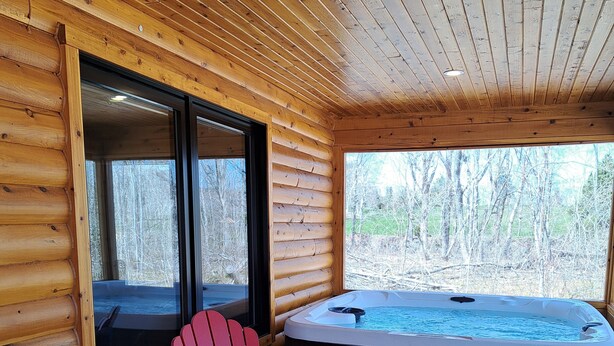 Interieur van een houten hutje met een hottub, een rode Adirondack-stoel en een raam met uitzicht op een bos.