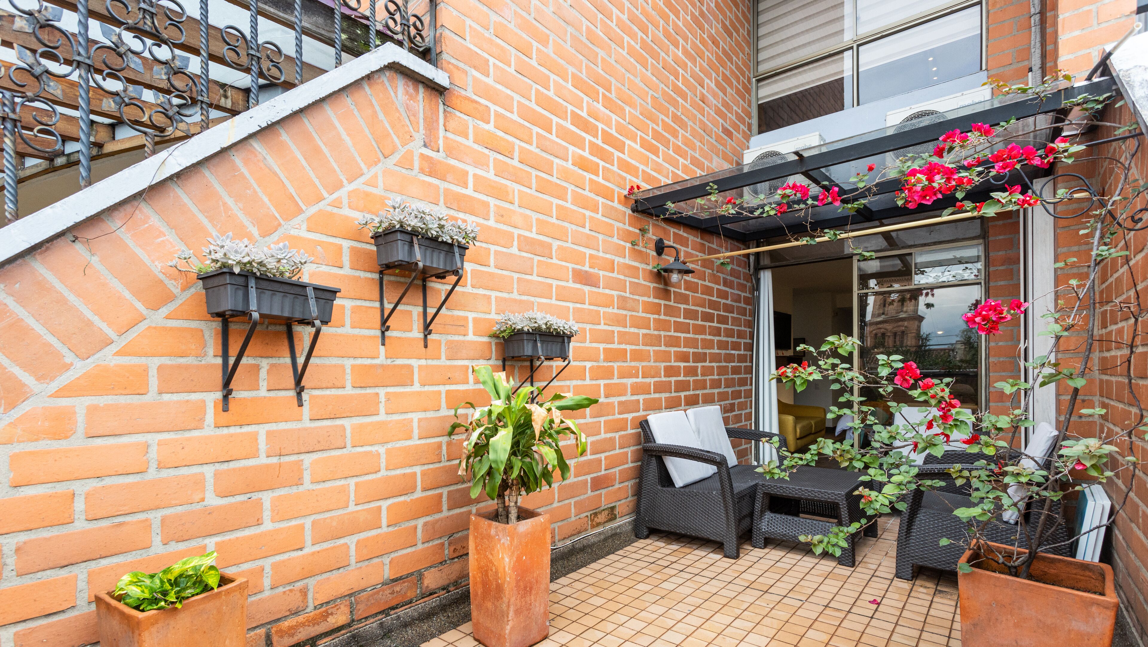 A brick exterior with outdoor seating, potted plants, and a pergola with flowers.