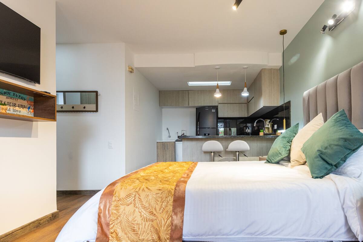 A modern bedroom with a large bed, a bedside table, and a view into the kitchen area.