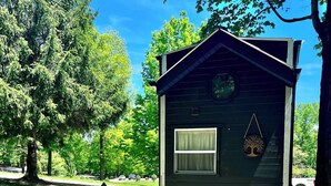 A small cabin with a window and a tree hanging ornament, surrounded by a gravel area with two lounge chairs and a small table. - Tiny House - Big Fun! (Zanesfield)