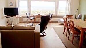 A living room with a sofa, a TV, a dining table with chairs, and large windows overlooking a scenic view. - Apartment in Nieuwpoort With sea View (Nieuwpoort)