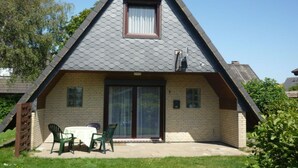 A small, single-story house with a triangular roof, a sliding glass door, and a small patio with a table and chairs. - North Sea Holiday House Wellmeyer (Wurster Nordseeküste)