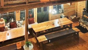 L’intérieur d’un chalet rustique en bois, avec de nombreuses bancs et tables en bois, une cheminée et une vue sur l’extérieur grâce à de grandes fenêtres.