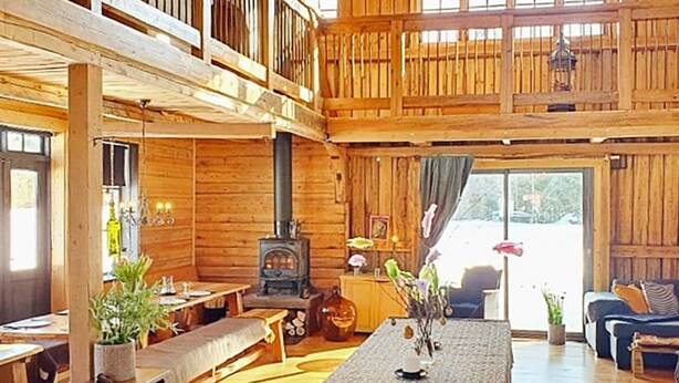 L’intérieur d’un chalet rustique, avec un plafond haut, des murs en bois, une cheminée centrale et un coin repas équipé d’une banquette.