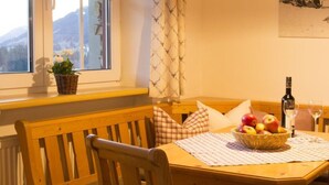 A cozy dining area with a wooden table, a basket of fruit, a bottle of wine, and a view of mountains through a window. - 2 in Alpenrosle Modern Retreat (Balderschwang)