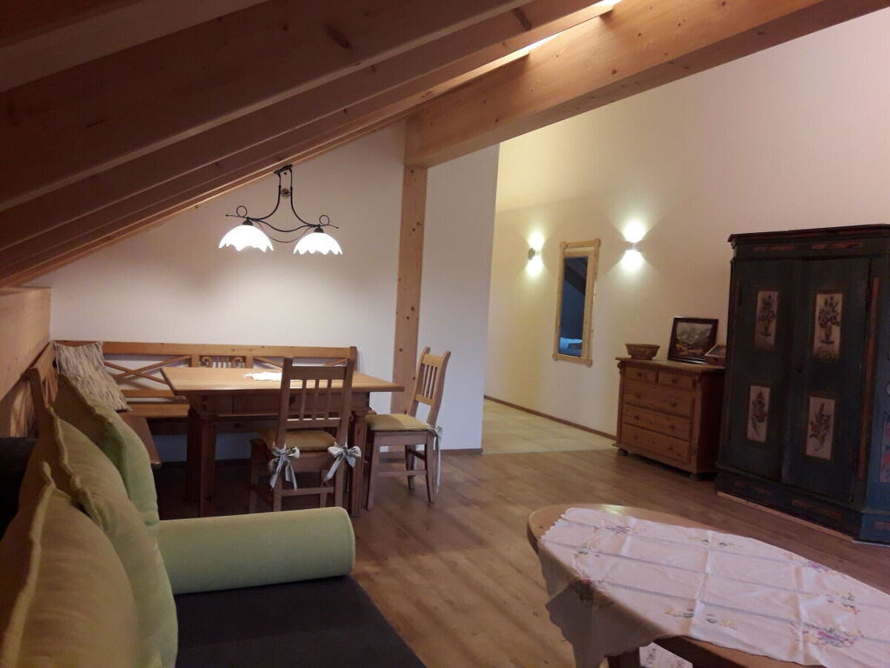 A cozy attic room with a wooden dining table, two chairs, a sofa, a small table with a tablecloth, and a cabinet with framed pictures.