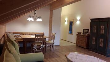 A cozy attic room with a wooden dining table, two chairs, a sofa, a small table with a tablecloth, and a cabinet with framed pictures.