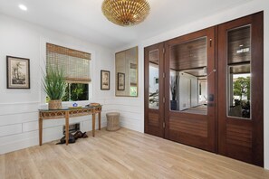Una habitación con piso de madera, una mesa consola de vidrio con una planta, un espejo grande y una puerta de madera con un ventanal.