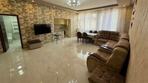A spacious living room with a large sofa, a dining table with chairs, a TV mounted on the wall, and a kitchen area visible through an open door.