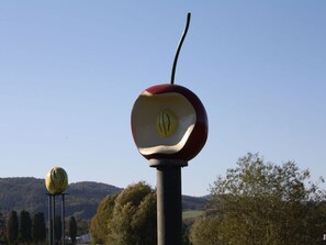 A sculpture of a red and yellow apple with a stem, set against a clear sky and a hilly landscape.