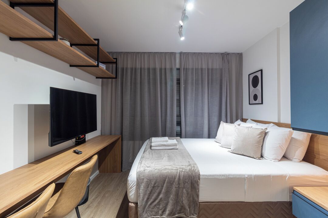Una camera d'albergo moderna con un letto, una scrivania con un computer, una televisione e un'ampia finestra con tende.