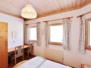 Ett rum med synliga träbjälkar i taket, en säng med ett vitt täcke, ett skrivbord med en stol och två fönster med blommiga gardiner.