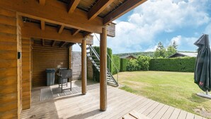 Une terrasse en bois avec un barbecue et des meubles d’extérieur, un patio couvert et une vue sur une pelouse avec une haie et des arbres.