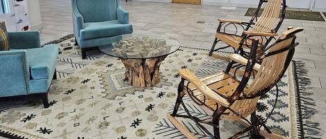 Un hall d’entrée avec un canapé bleu, un fauteuil à bascule en bois et une table basse en verre. Un tapis à motifs recouvre le sol, une porte se trouve au fond et une petite table avec une plante est également présente.