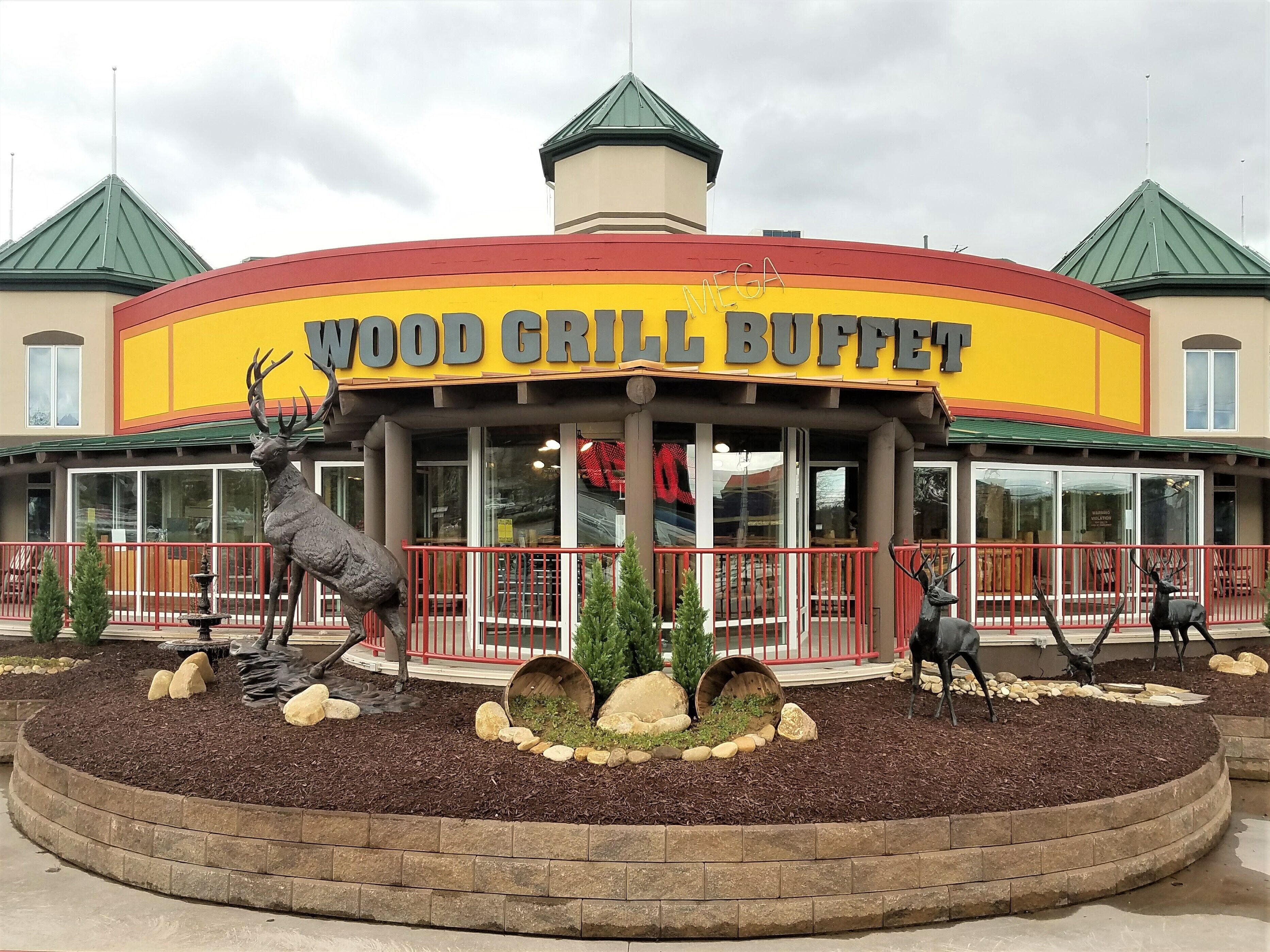 Ein Restaurant namens "Wood Grill Buffet" mit einer markanten roten und gelben Fassade, einem zentralen Turm und Gartenskulpturen von Hirschen.