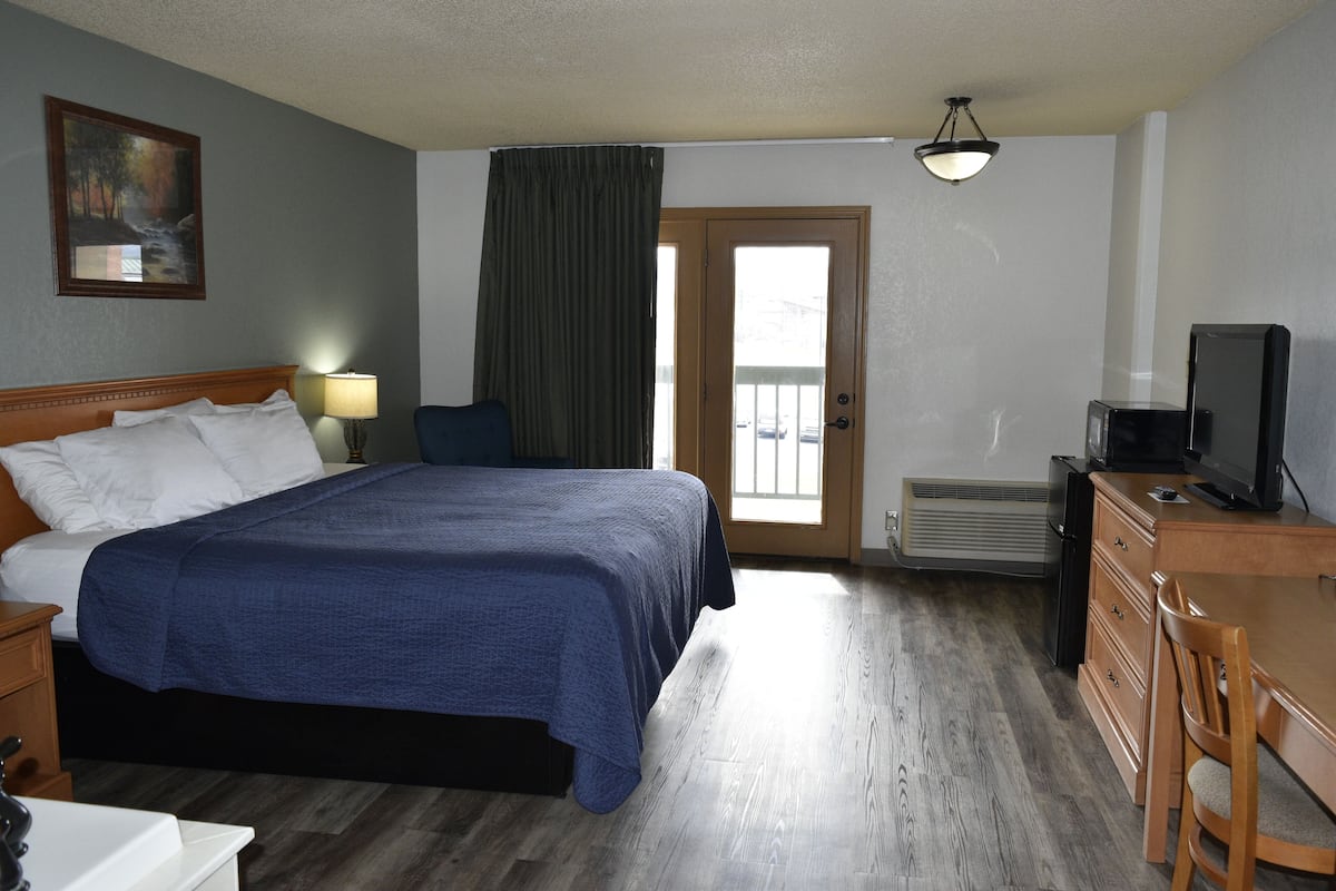 Une chambre d’hôtel avec un grand lit, un bureau avec une télévision, une commode, une fenêtre avec des rideaux et un tableau au mur.
