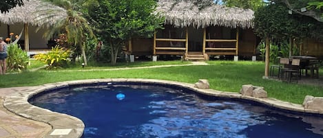 Una piscina con borde de piedra, rodeada de césped y árboles. Se observan estructuras con techo de paja al fondo.