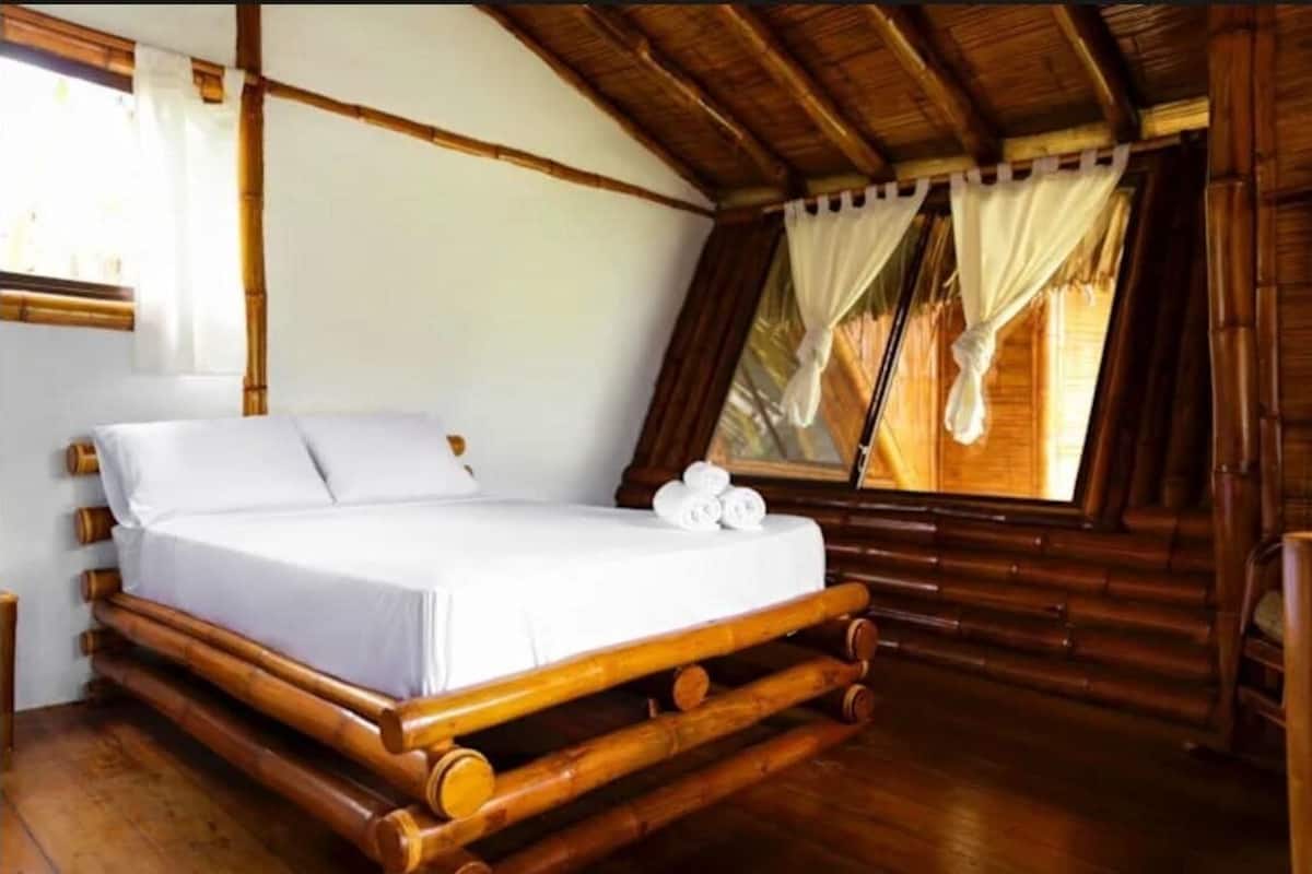 Una habitación con estructura de madera, una cama, un ventanal con cortinas y suelo de madera.