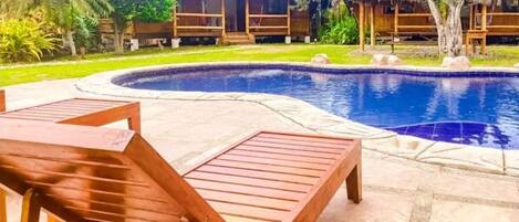 A swimming pool with wooden loungers and thatched-roof structures in the background.