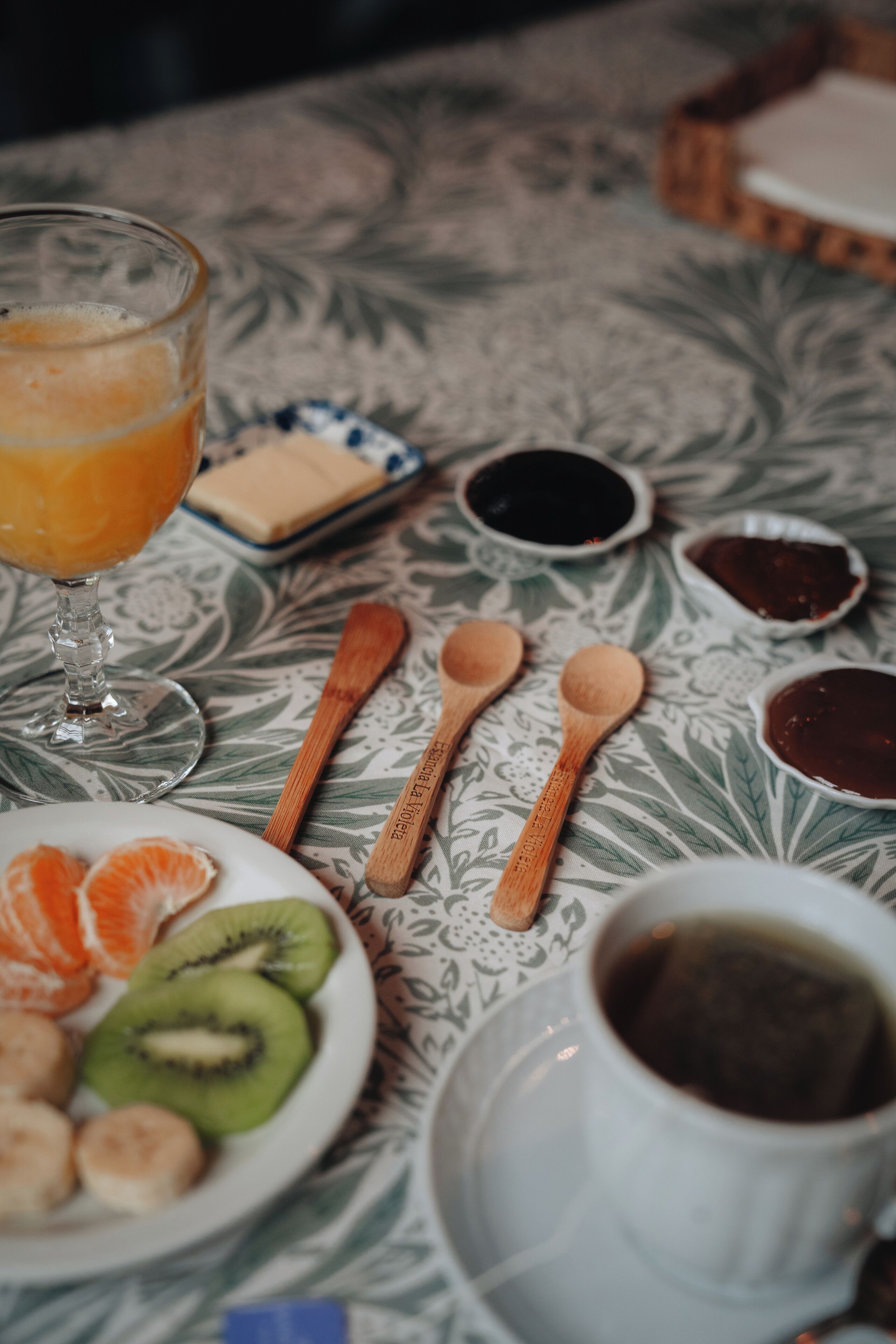 A table with a floral tablecloth, a glass of orange juice, a plate with sliced kiwi and oranges, wooden spoons, and small bowls of condiments.