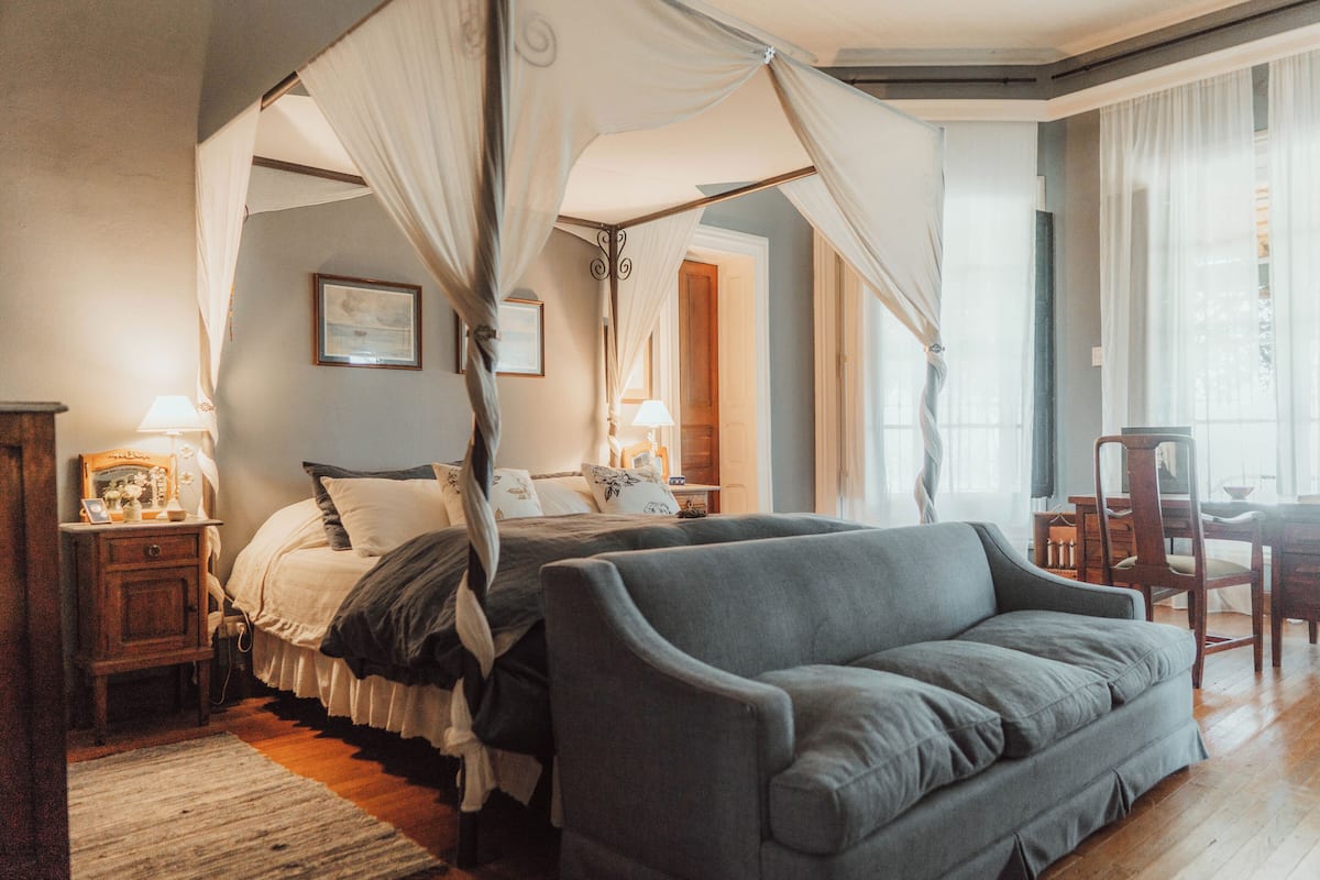 Quarto com cama de dossel, um sofá cinzento, mobiliário de madeira e uma janela grande.