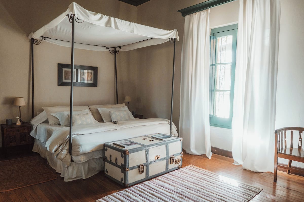 Uma cama de dossel com estrutura de madeira, uma mesinha de cabeceira com abajur, uma mala e uma janela com cortinas.