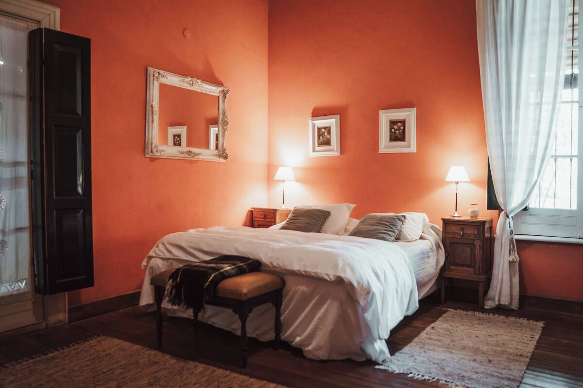 Quarto com cama, mesas de cabeceira, espelho e janela com cortinas.