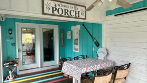 A turquoise-walled porch with a ceiling fan, a table with a colorful tablecloth, and two wicker chairs.