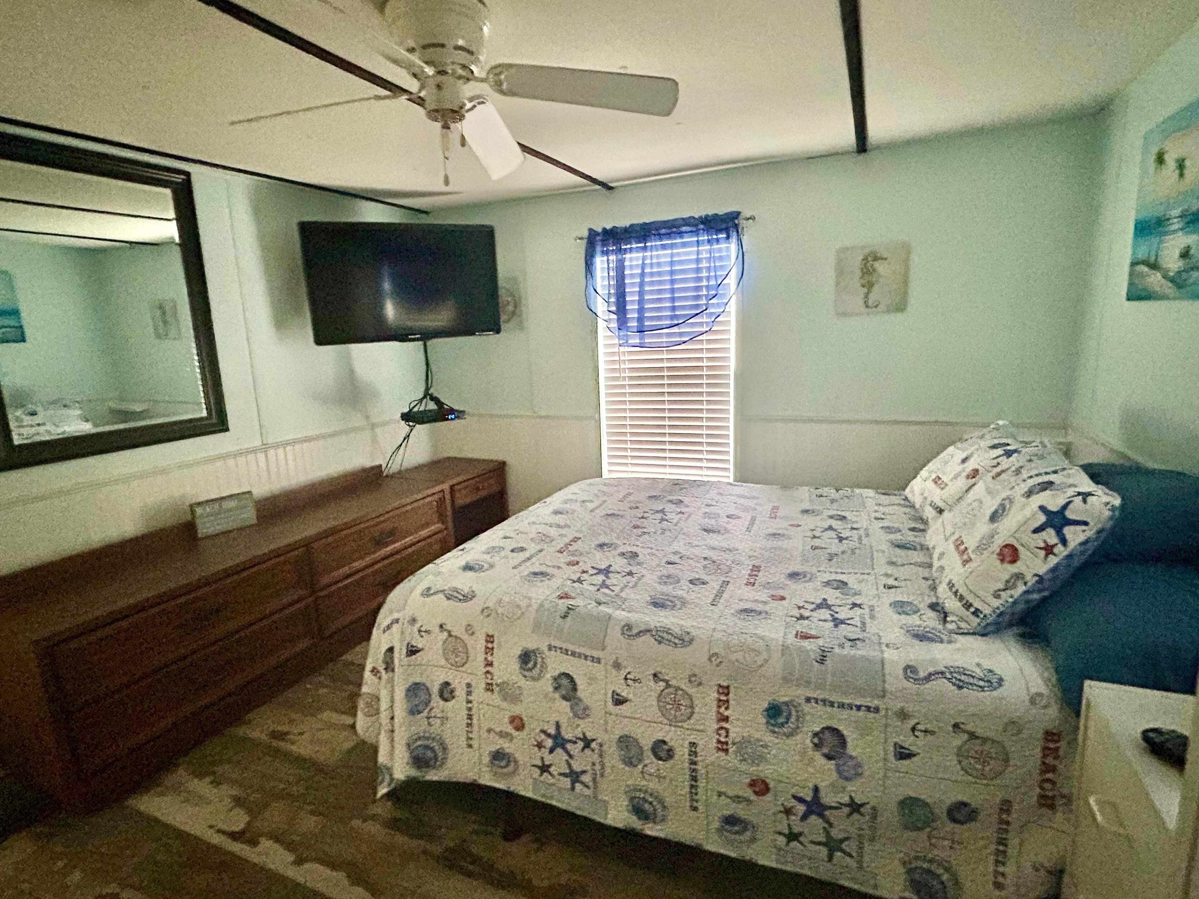 Un dormitorio con cama, cómoda, televisor, ventilador de techo y ventana con persianas.