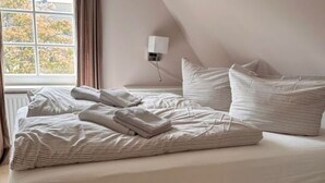 Una cama bien hecha con almohadas y toallas dobladas en una habitación con techo inclinado y una ventana con cortinas.