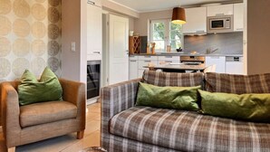 Una sala de estar moderna con un sofá y un sillón, una cocina con armarios blancos y un microondas, y un papel tapiz estampado en las paredes.