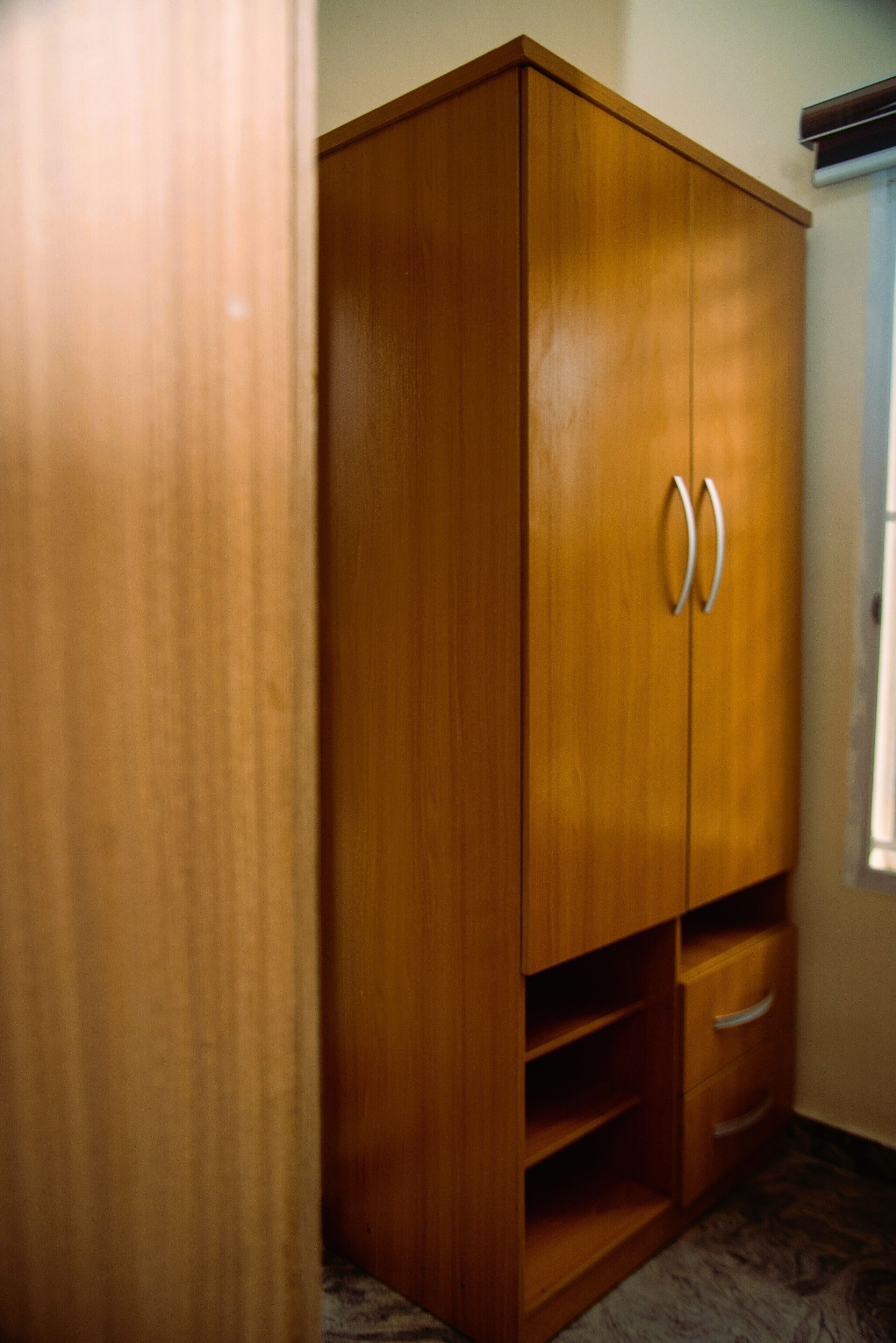 Une armoire en bois à deux portes et une étagère, située dans une pièce dont le sol est recouvert d’un tapis à motifs.