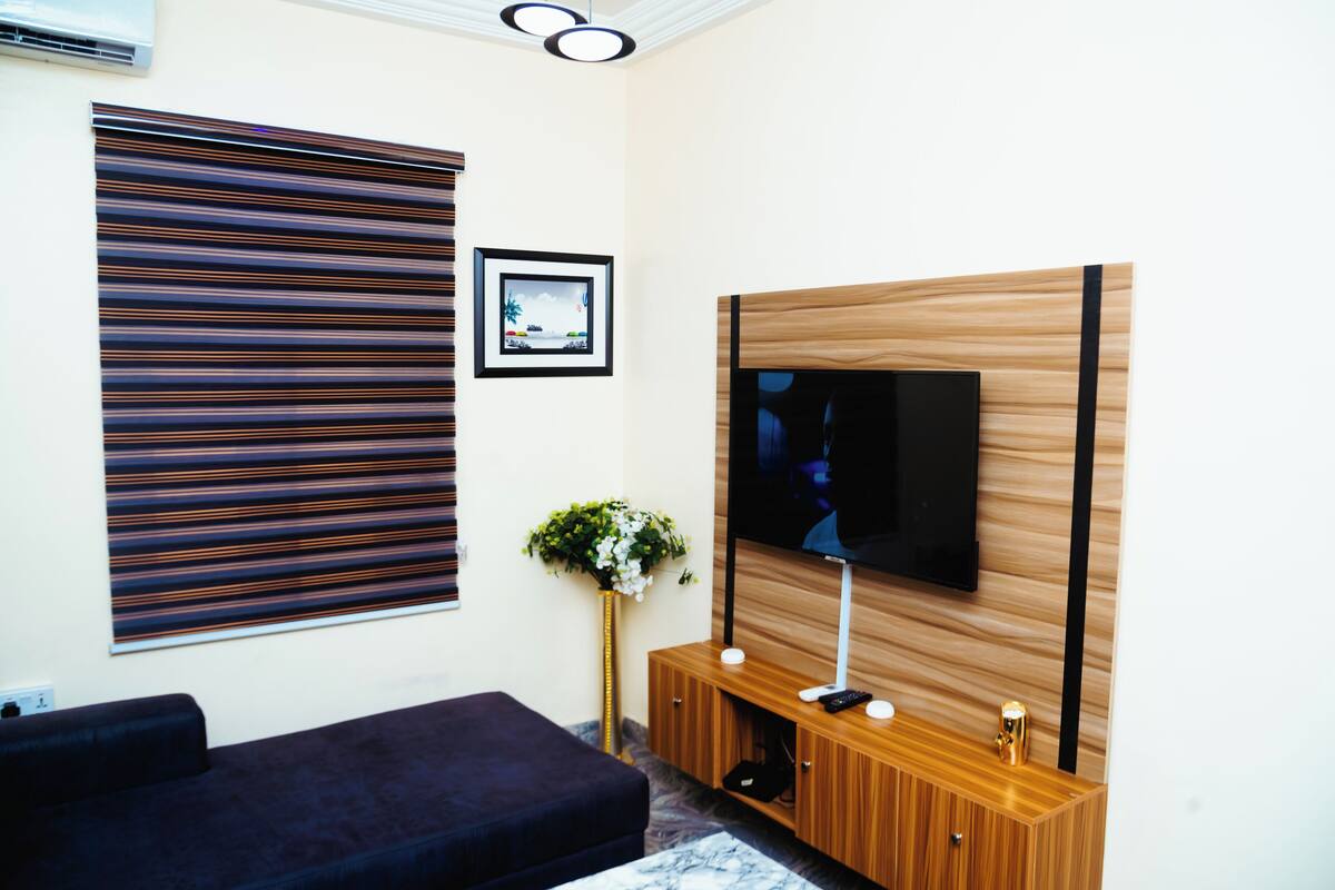 Ein modernes Wohnzimmer mit einem dunkelgrauen Sofa, einer Holz-Unterhaltungseinheit mit einem Flachbildfernseher, einem großen, gestreiften Fensterladen und einem gerahmten Bild an der Wand.
