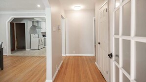 A hallway with wooden flooring, white walls, and a kitchen area visible through an open door.