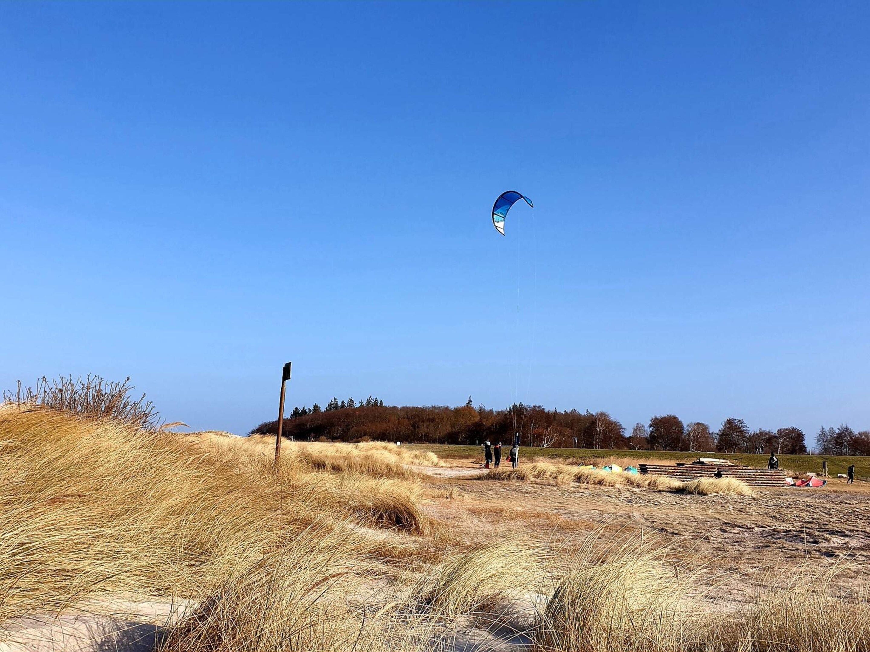 A clear sky with a kite flying. There are dry grass fields and a few people in the distance.