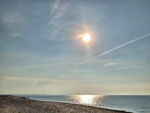 Ein Strand mit klarem Himmel, der Sonne tief am Horizont und einem fernen Leuchtturm.