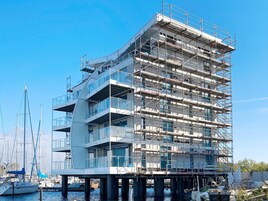 A multi-story building with a modern architectural design, featuring balconies and large windows, situated on stilts over water with boats in the background.