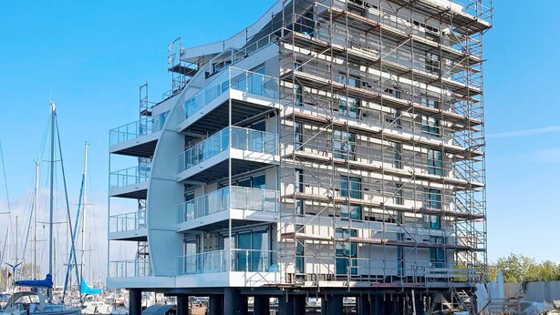 Ein mehrgeschossiges Gebäude mit moderner Architektur, das Balkone und große Fenster aufweist und auf Stelzen über dem Wasser steht, mit Booten im Hintergrund.