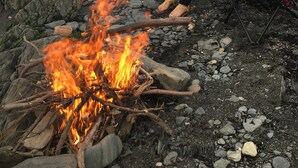 Two people sitting on chairs by a campfire on a rocky beach.
