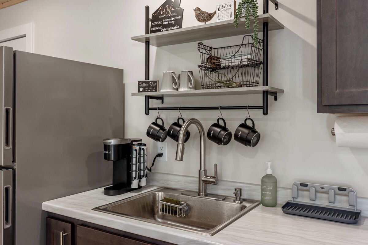 Eine Küche mit einem Edelstahlkühlschrank, einer Kaffeemaschine, einem Wasserkocher und einem Wasserkocher-Ständer. Es gibt ein Regal mit einer Vogel-Figur, einem Schild mit der Aufschrift 'Coffee Bar' und einer Pflanze.