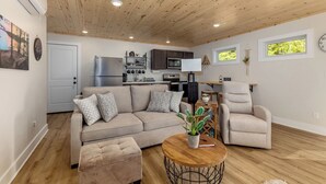 Una sala de estar moderna con techo de madera, un sofá, una silla, una mesita redonda y una cocina al fondo.